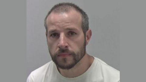 Police mugshot of a man with cropped dark hair and a short beard and moustache against a grey background.