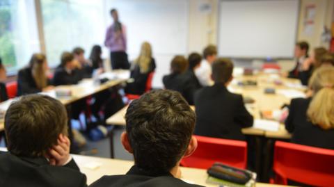 A stock image of pupils sitting at their desks in a classroom. The shot is taken from the back of the room and none of the children's faces is visible or recognisable. The back of the heads of two boys are in the foreground in focus, while the rest of the children in the room and the teacher standing at the front are out of focus. All the children are in uniform, wearing dark blazers.