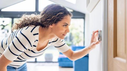 A woman dressed in a striped short-sleeved shirt and jeans adjusts a wall-mounted thermostat by her door. She is in a light-filled room with large windows
