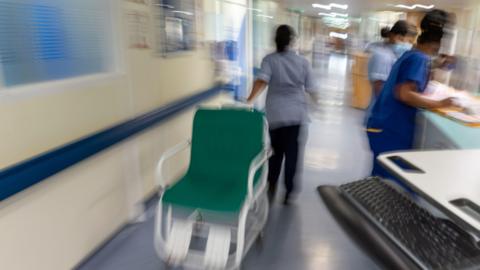 A busy ward in a hospital. One member of staff in uniform is pulling a green chair. The background is blurred.