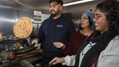 A young man flips a paratha in a frying pan while two women with floured hands help