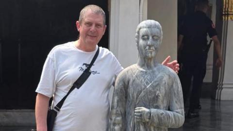 Paul Lynn in a white T-shirt with a small black bag over his shoulder. He is posing next to a grey statue of a priest-like figure. There are marble floors with pillars that have gold tops. There is a security guard in the background. 