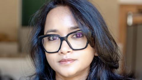 A woman - Prof Ronita Bardhan - looking directly at the camera with a neutral expression. She is wearing black glasses and has dark hair.