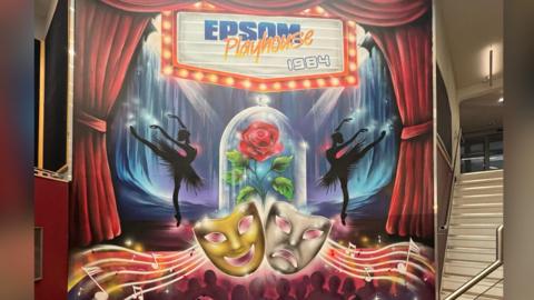 A mural of a stage. The stage has blue and purple lights, two silhouettes of ballerinas and a glass with a red rose inside. At the bottom of the mural is musical notes joined with a gold and silver mask. 