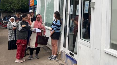 People queue on a pavement outside a building.