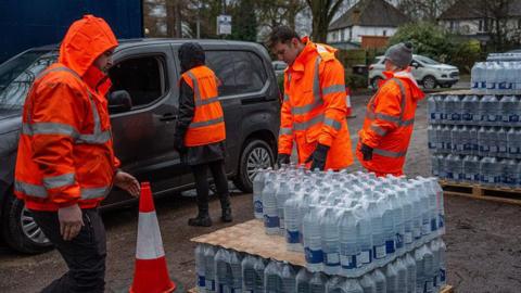 Four people in hi-vis orange jackets taking out bottled water supplies out of vehicles at a carpark.