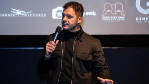 Joshua Ford speaking on stage. He holds a microphone and wears a black top and black jacket. 