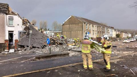 A scene showing the destroyed house, which has been flattened alongside a row of houses. In the foreground, two people wearing fire service uniforms are seen