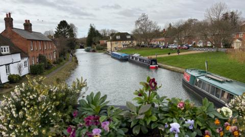 A canal runs through the centre of the picture with three barges on the right-hand side. In the foreground there is a flower box full of brightly coloured flowers and beyond the barges are stretches of green grass lined by cars and houses. There are also a few houses on the left-hand side of the canal.