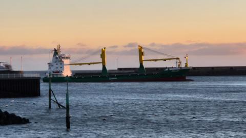 A large cargo ship which is white, blue and yellow. The front of the ship is slightly higher in the water than the back. The sea looks calm and there is a long pier behind it and what looks like another ship to the left. The sun is still rising and the sky is blue and yellow.