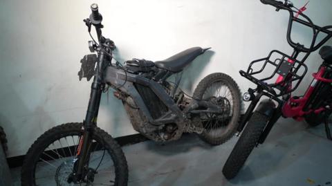 A grey motorbike leaning against a white wall.  It's quite muddy, as if it's been ridden off road.