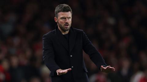 Manchester United interim head coach Michael Carrick makes a palms-down gesture that appears to be calling for calm as he wears a dark coat over a dark top