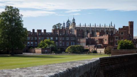 Eton College seen from a distance. It's a large red brick building with multiple spires and turrets, with a well-kept lawn next to it.