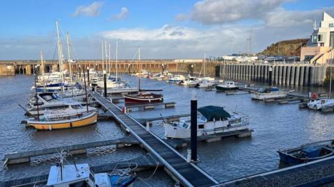 Watchet harbour at high tide, with various types of boats floating on the water. It's sunny but there are also some clouds in the sky