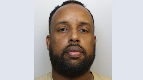 A mugshot of Mohamed Osman. He is wearing a yellow t-shirt and standing against a grey background. He has dark hair and a goatee style beard. 