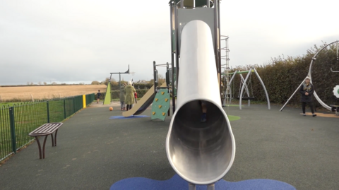 A new play area including a metal slide, rubber flooring and a range of apparatus. People can be seen playing and grey skies behind.