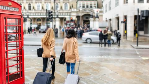 Two women wearing tan coats walk along a wet London street, each pulling a suitcase, beside a red telephone box with traffic and pedestrians in the background.