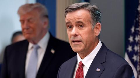 Ratcliffe looks at the camera while speaking with Trump out-of-focus in the background next to a US flag, pictured in the White House in April.