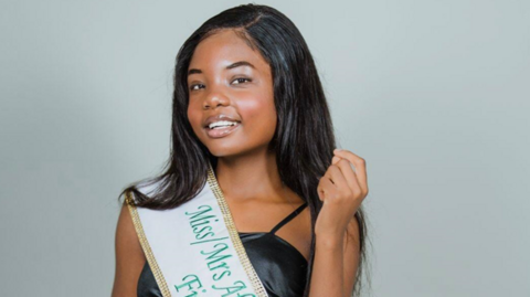 Shows a young woman with long black hair and a black top with a green and white sash 