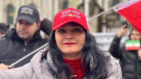A woman with black hair is standing outside at a protest in front of a building. She is wearing a red hat, top and grey coat. Other people can be seen wearing coats behind her. A small Iranian flag can be seen held by someone out of focus behind her.