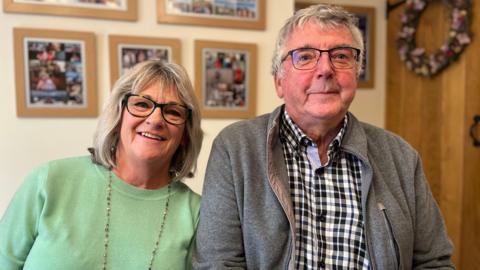 Kathy and Steve smile. They sit with blurred family photos behind them. Both wear glasses. Kathy on the left wears a mint green jumper while Steve has a check shirt and grey fleece.