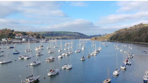 An estuary full of small sailing boats with hills on either side and blue cloudy sky above