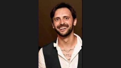 Steven Morris wearing an open necked white shirt and a black waistcoat. He has short brown hair and a brown beard.