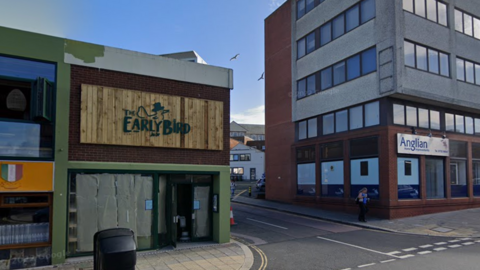 A Google street view image of the Early Bird cafe. It is a green building with the bird and name on the logo. There is a road to the right of it and a building to the right of the road. The sky is blue. There is a bin in front of the cafe. 