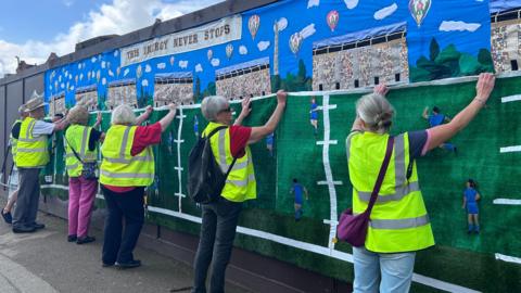 Five people dressed in high-visibility jackets hold up a large knitted mural and place it on a charcoal wall.