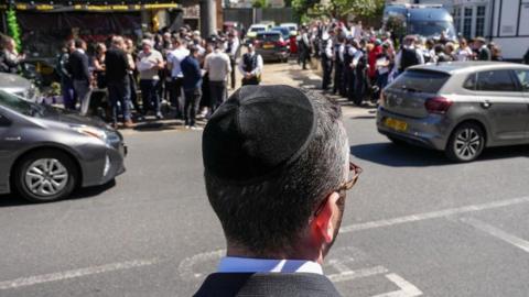 A Jewish man wearing a kippah is seen from behind as he stands looking out at a road as two cars pass in Golders Green. On the opposite pavement crowds are gathered with police officers nearby.