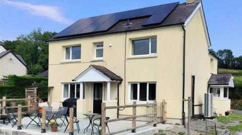 A two-storey cream-coloured house with a pitched roof fitted with large black solar panels. The house has several white-framed windows, a small covered front porch, and an elevated concrete patio area with outdoor tables, chairs, and wooden railing posts.