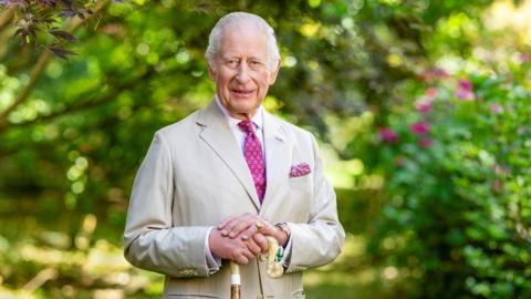 King Charles in a pale beige linen suit with a cane in front of a garden