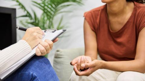 Close-up of an unrecognisable patient talking to a lady who appears to be making notes on a clipboard.