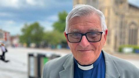 The head and shoulders of Reverend Albert Cadmore, who wears a grey suit jacket, blue shirt and white dog collar. He has black-framed glasses. He looks at the camera. Out of focus behind him is a pedestrianised area, a church and trees.