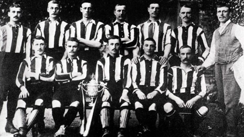 A black and white image of footballers in striped kit, seated in two rows, with a man in a suit to the righthand side of the image.

On the lower row, an ornate cup is perched on the edge of a chair between two of the footballers.