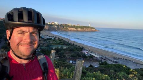A man wearing a helmet stands looking at the camera. A scenic view of the sea, beach and a lighthouse can be seen in the background behind him.