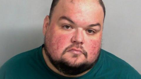 Police custody image of Gavin Plumb. He has receding hair cut short, a beard and moustache, and is wearing a turquoise top/