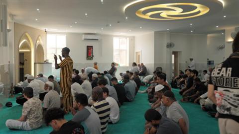 The inside of the mosque - a bright room with green carpet. A large group of men sit on the carpet