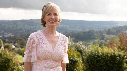 Kate Turner is standing on a hill in a pink dress. She is smiling at the camera as the sun shines on her. She is in a pink lacey and floral dress and has bob-length blonde hair. There is greenery and country hills behind her.