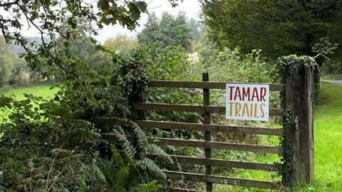 A wooden gate next to a hedge and field with a sign that reads Tamar Trails 