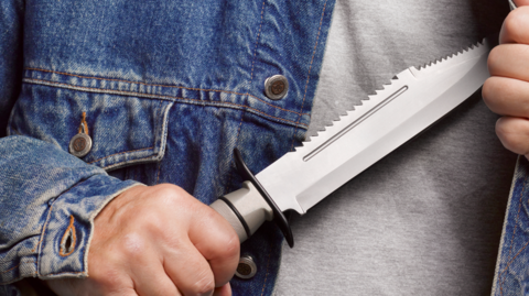 A hand drawing a serrated knife from inside a denim jacket