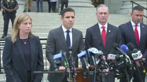 Prosecutor wearing a black suit speaks into mic, with three people around him.