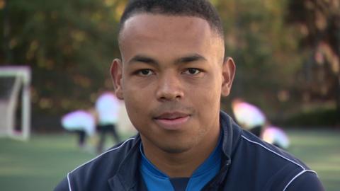 Close-up of Johnny Byamungu who is seen above the shoulders smiling at the camera. He is wearing a tracksuit jacket and children playing on astroturf can be seen in the background.