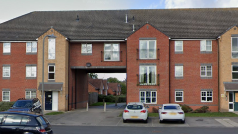 A three storey residential block with an archway underneath part of the building. Four cars are parked outside the front of the building.