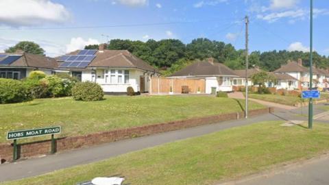 A sign for Hobs Moat Road can be seen on the pavement of a grassy verge. A umber of bungalows and two storey homes are pictured in the distance, with a grass lawn behind the road sign.