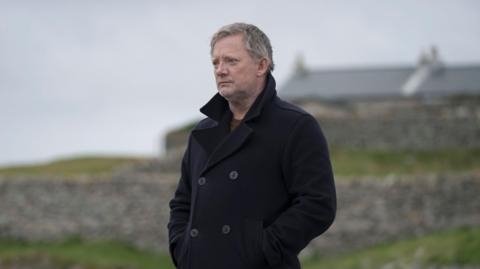 Douglas Henshall in charcter as DI Jimmy Perez, he is wearing a black coat and is looking into the distance