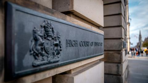 A general view outside the High Court in Edinburgh. A black sign with dark lettering and a coat of arms is attached to light stone bricks. The lettering reads "High Court of Justiciary". 