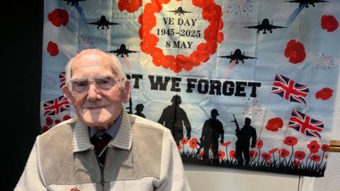 An old man with glasses, looks at the camera, as he sits in front of large poster. The poster has a blue sky background with red poppies on it and silhoutees of soldiers, with British flags. It reads 'lest we forget' and 'VE DAY 1945-2025- 8 MAY'