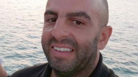 Samir Zitouni is smiling at the camera in a close-up shot of his face. He is balding and has a moustache and a beard