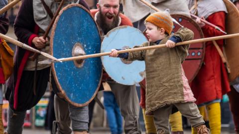 A little boy in costume wields a spear while men behind him hold large wooden shields
